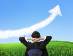 A man in a business suit leans back in an office chair with his hands behind his neck. He is looking at a field of lush green grass and a blue sky filled with a cloud shaped like an arrow pointing up.