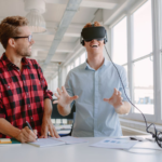 A young man wearing a plaid flannel shirt watches his coworker use a virtual reality headset to explore a video game scene.