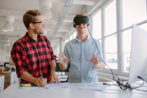 A young man wearing  a plaid flannel shirt watches as his coworker uses a virtual reality headset to explore an artificial setting.
